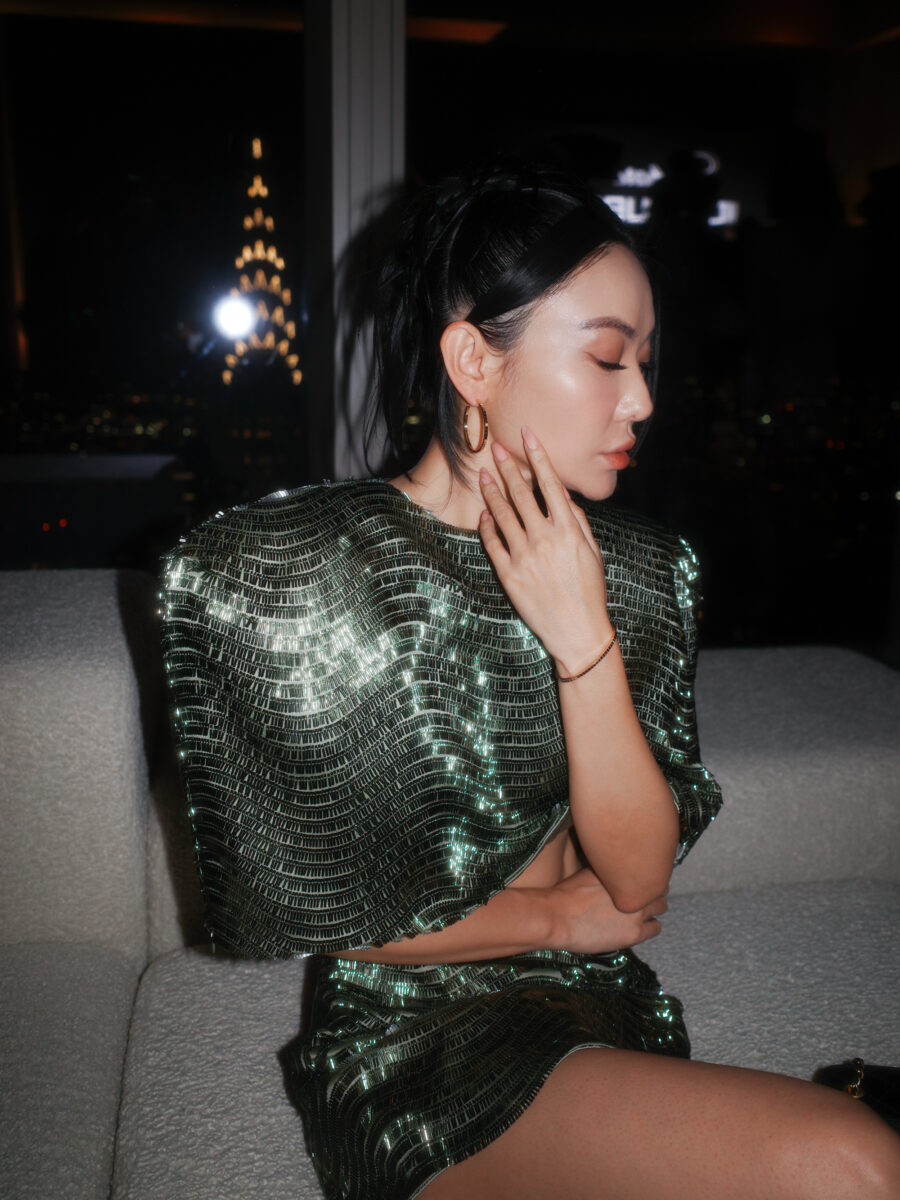 Jessica Wang in a shimmering green evening look touching her neck, photographed against the New York skyline at night.