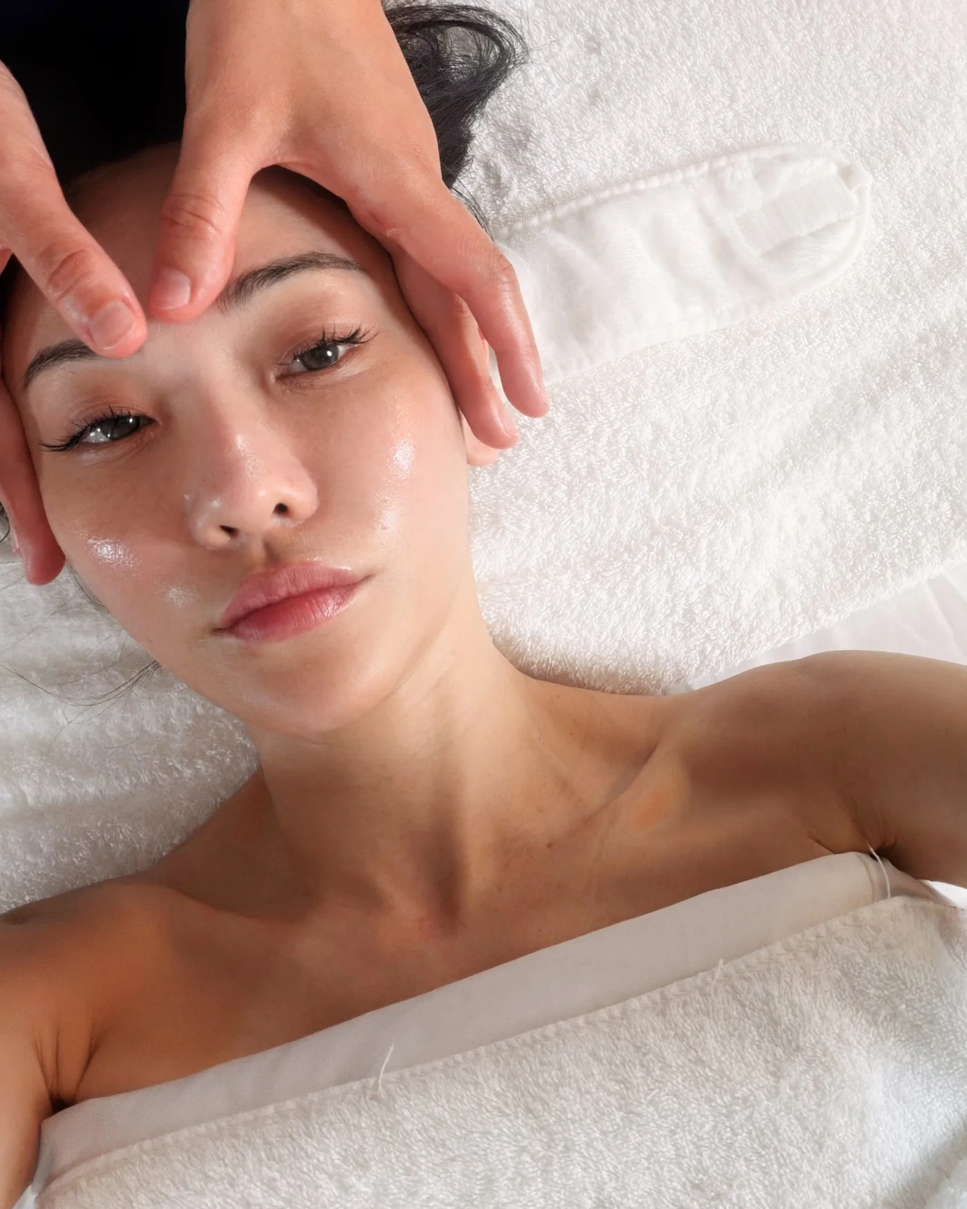 Jessica Wang reclines under white towels while another person's hands frame her forehead and cheeks.