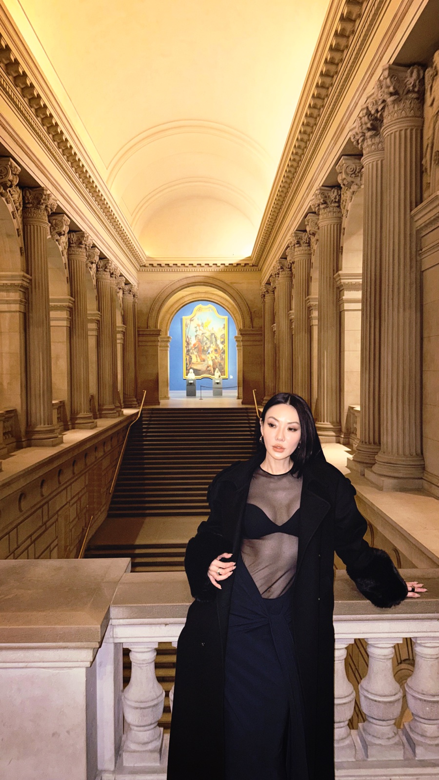 Jessica Wang wearing a sheer black top and long black coat in a columned interior